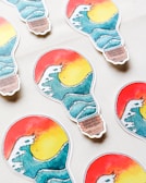 Several stickers shaped like light bulbs are arranged on a light surface. Each sticker features an artistic design of a wave overlaid on a sunset with vibrant colors. The top half showcases a red and yellow gradient resembling a sunset, while the bottom half displays a stylized blue wave.