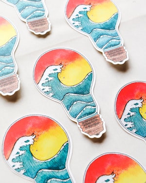 Several stickers shaped like light bulbs are arranged on a light surface. Each sticker features an artistic design of a wave overlaid on a sunset with vibrant colors. The top half showcases a red and yellow gradient resembling a sunset, while the bottom half displays a stylized blue wave.