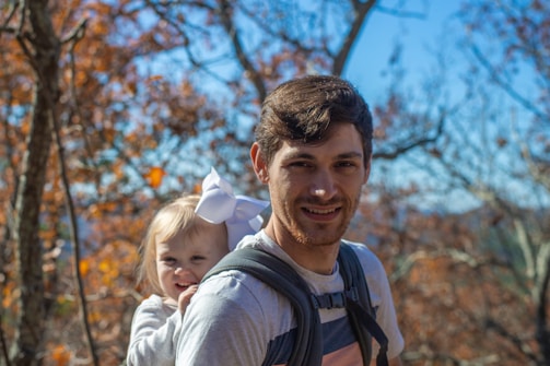 A man with short dark hair is carrying a smiling child with a large white bow in her hair on his back using a baby carrier. The background consists of trees with autumn leaves and a clear blue sky.