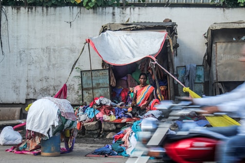 A person sits under a makeshift shelter made of tarpaulin, surrounded by a large collection of colorful clothes. The clothes are piled on the ground and draped over a drum. A blurred motorcycle passes by in the foreground, suggesting motion and busy street life.