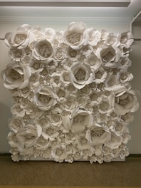A wall covered with large, intricately crafted paper flowers in various sizes, predominantly in shades of white. The flowers are arranged densely, creating a textured, artistic backdrop. The setting appears to be indoors with soft overhead lighting illuminating the display.