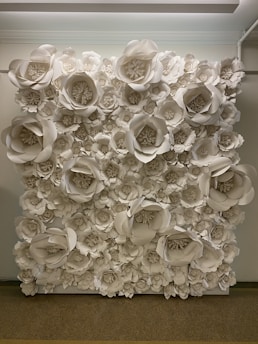 A wall covered with large, intricately crafted paper flowers in various sizes, predominantly in shades of white. The flowers are arranged densely, creating a textured, artistic backdrop. The setting appears to be indoors with soft overhead lighting illuminating the display.