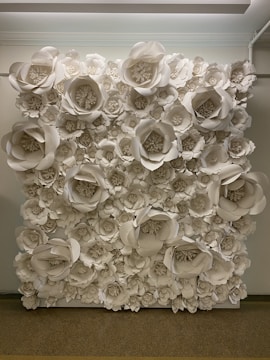 A wall covered with large, intricately crafted paper flowers in various sizes, predominantly in shades of white. The flowers are arranged densely, creating a textured, artistic backdrop. The setting appears to be indoors with soft overhead lighting illuminating the display.