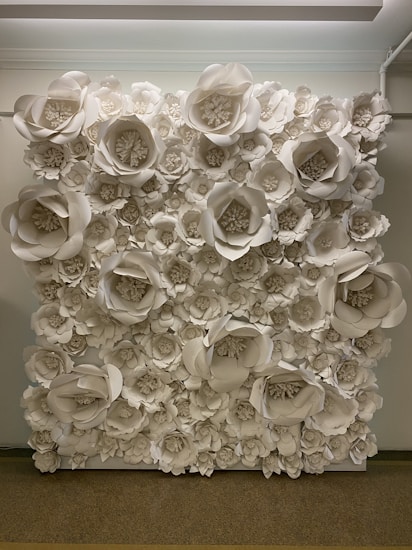 A wall covered with large, intricately crafted paper flowers in various sizes, predominantly in shades of white. The flowers are arranged densely, creating a textured, artistic backdrop. The setting appears to be indoors with soft overhead lighting illuminating the display.