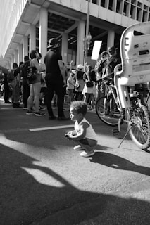 A black and white photograph of a young child crouching on a city street, among a group of people who are participating in a public gathering or protest. A bicycle with a child seat is nearby, and several individuals are holding signs. The setting appears to be urban, with a backdrop of modern architecture.