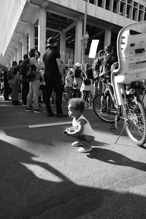 A black and white photograph of a young child crouching on a city street, among a group of people who are participating in a public gathering or protest. A bicycle with a child seat is nearby, and several individuals are holding signs. The setting appears to be urban, with a backdrop of modern architecture.