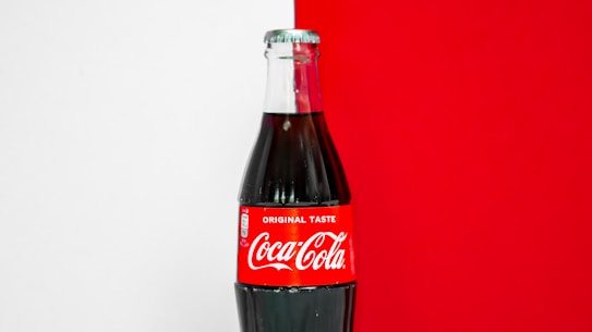 A classic glass bottle of Coca-Cola with a red label that prominently displays the brand's name in white lettering. The bottle is against a background that is half white and half bright red.