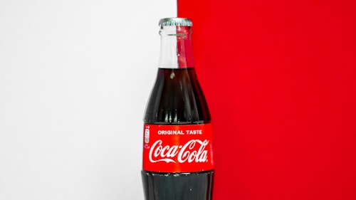 A classic glass bottle of Coca-Cola with a red label that prominently displays the brand's name in white lettering. The bottle is against a background that is half white and half bright red.