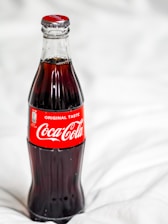 A classic glass bottle filled with Coca-Cola, featuring the iconic red label with white text. The bottle is placed against a soft, white, blurred background, highlighting its dark contents and slender shape.
