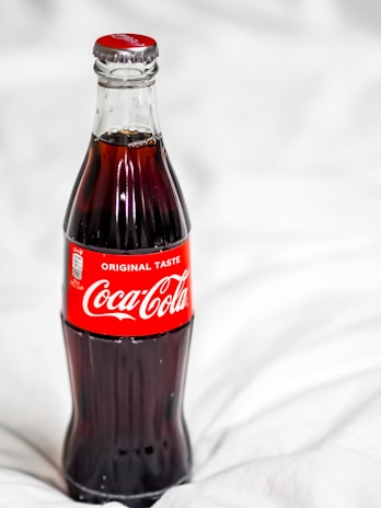 A classic glass bottle filled with Coca-Cola, featuring the iconic red label with white text. The bottle is placed against a soft, white, blurred background, highlighting its dark contents and slender shape.
