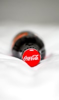 A close-up of a bottle lying on a surface, focused on the red cap with the Coca-Cola logo. The background is out of focus, emphasizing the brand prominently.
