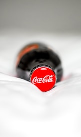 A close-up of a bottle lying on a surface, focused on the red cap with the Coca-Cola logo. The background is out of focus, emphasizing the brand prominently.
