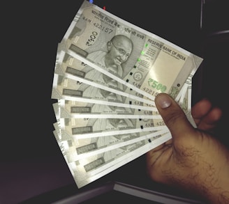 A hand holding multiple Indian 500 rupee banknotes, fanned out. The banknotes feature a portrait of Mahatma Gandhi and are from the Reserve Bank of India.