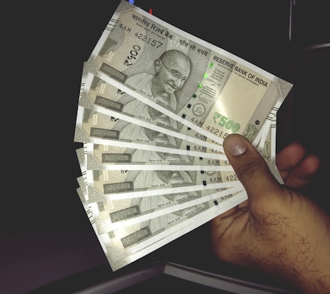 A hand holding multiple Indian 500 rupee banknotes, fanned out. The banknotes feature a portrait of Mahatma Gandhi and are from the Reserve Bank of India.