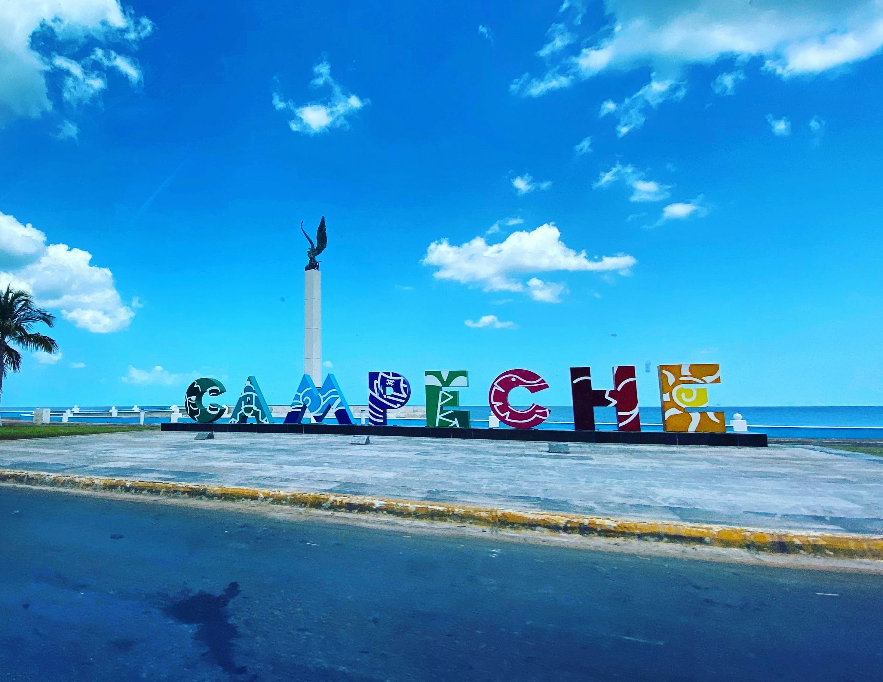 A colorful sign spelling out 'CAMPECHE' in large artistic letters is set against a scenic backdrop of the ocean and a vibrant blue sky. There is a monument with a statue nearby and a palm tree to the side, all adding to the picturesque coastal setting.