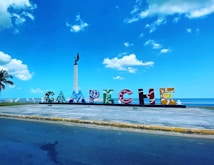 A colorful sign spelling out 'CAMPECHE' in large artistic letters is set against a scenic backdrop of the ocean and a vibrant blue sky. There is a monument with a statue nearby and a palm tree to the side, all adding to the picturesque coastal setting.