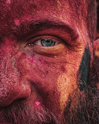 A close-up of a person's face covered in vibrant colored powders, focusing on the eye. The textures of the skin and powder create a rich, detailed pattern with various hues of red, pink, and yellow.