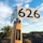 A large cricket bat stands upright with a sign displaying the number 626 affixed to its handle. The bat features a sticker from thecricketlibrary.com, promoting their love for cricket books. In the background, a palm tree and expansive shrubbery stretch under a vibrant blue sky with scattered clouds.