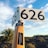 A large cricket bat stands upright with a sign displaying the number 626 affixed to its handle. The bat features a sticker from thecricketlibrary.com, promoting their love for cricket books. In the background, a palm tree and expansive shrubbery stretch under a vibrant blue sky with scattered clouds.