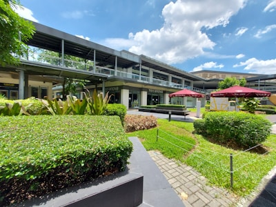 A modern, open-air shopping area with lush greenery, including bushes and trees. The foreground features neatly trimmed hedges and a pathway made of stone tiles. Several small umbrellas with seating areas are visible, along with a multi-level building displaying signs.