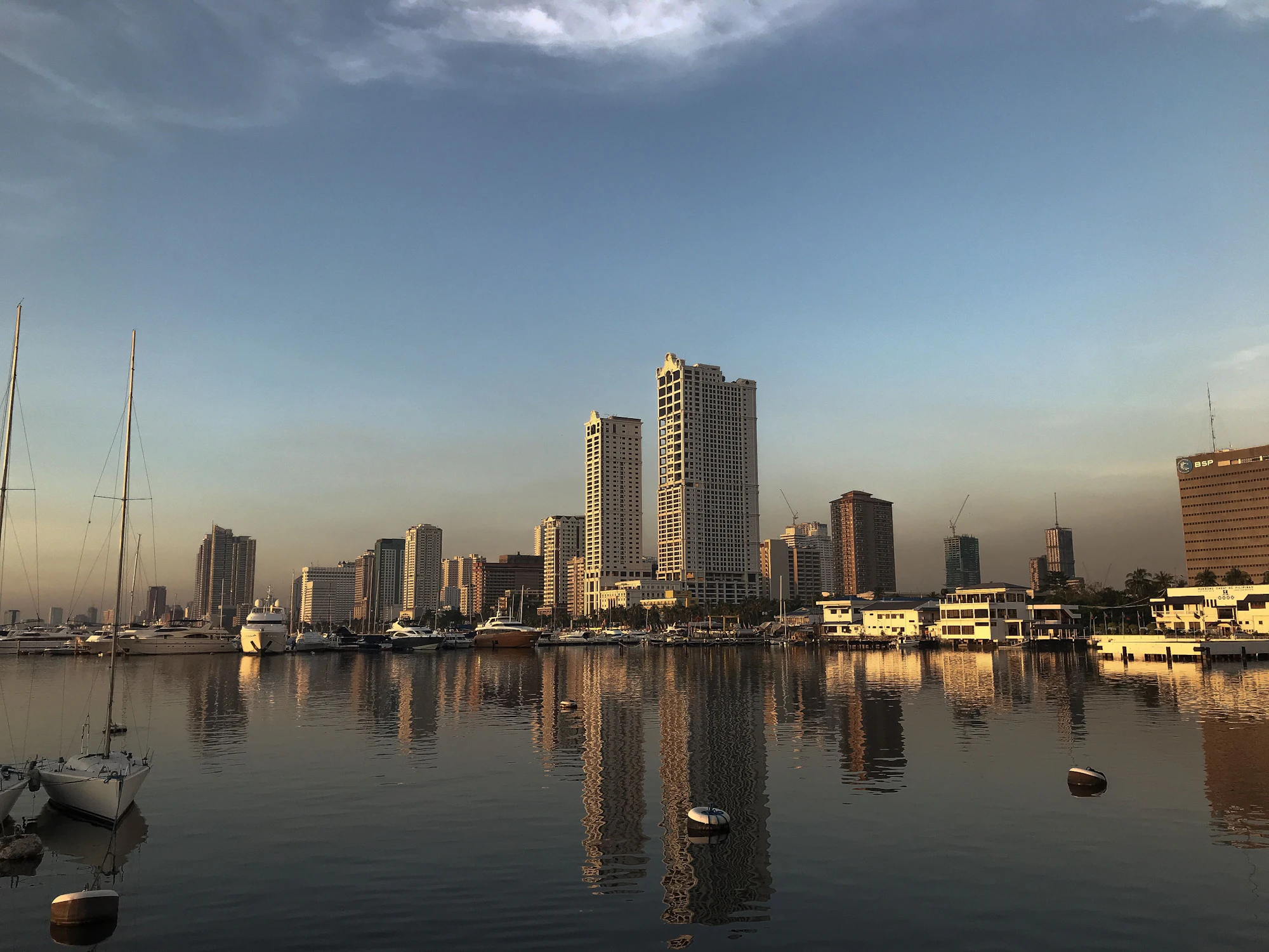 The Manila skyline across the bay at dusk, towers catching the last light with moored boats and their reflections in the water in front