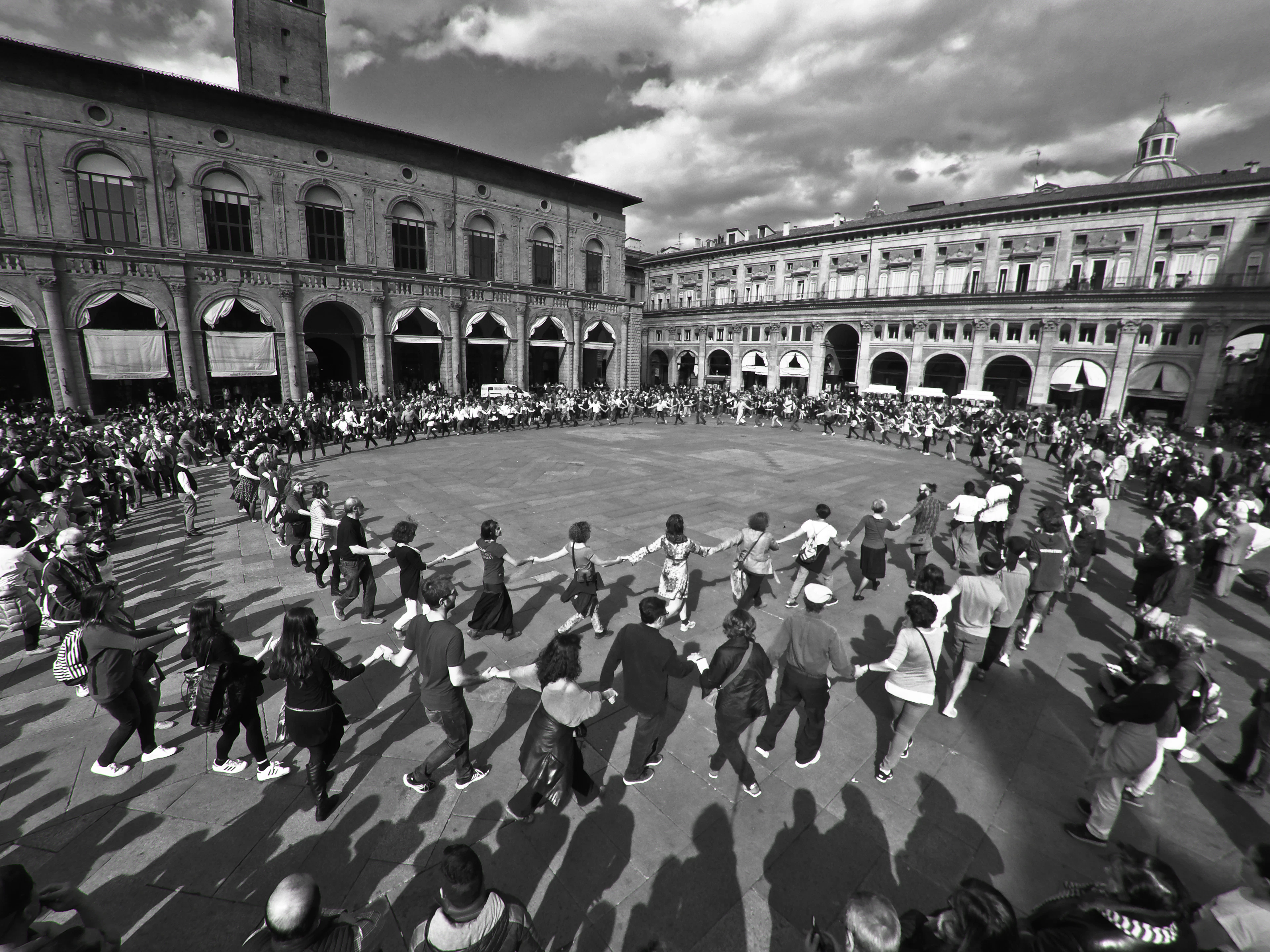 Black-and-white photo of a large crowd dancing in a circle in a historic Bologna piazza.