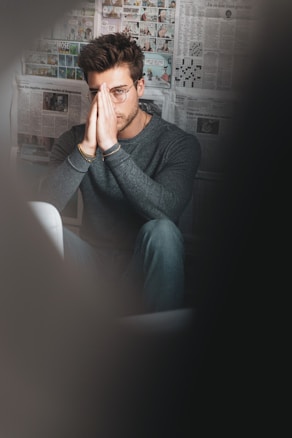 A man with glasses sitting indoors against a background covered in various newspaper clippings and illustrations. He is wearing a gray sweater and blue jeans, with his hands pressed together in front of his face in a contemplative or thoughtful pose.
