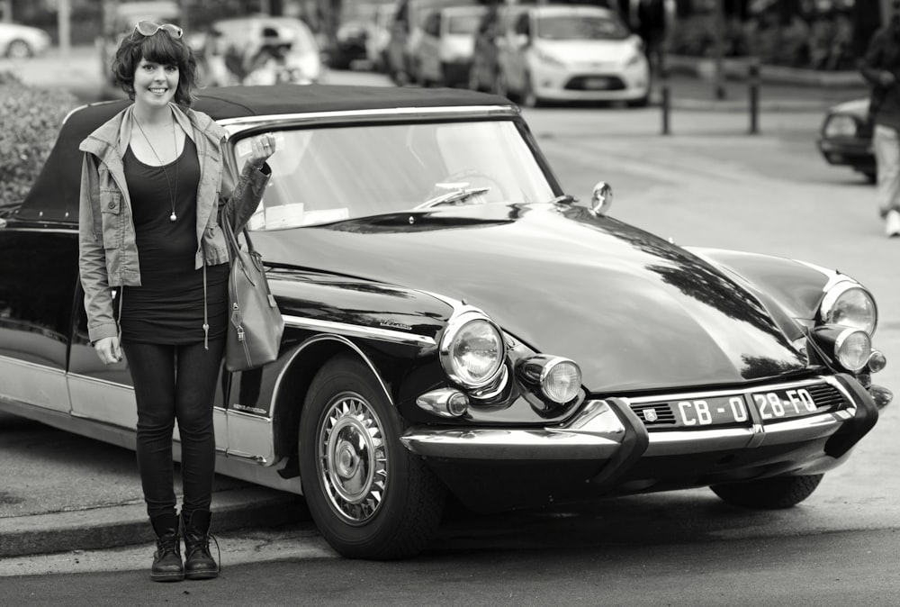 grayscale photo of woman in black sleeveless dress standing beside classic car