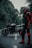 A motorcyclist in full gear, including a helmet and protective suit with red accents, stands near a sleek black sports motorcycle. The scene is set on a quiet, tree-lined road, conveying a sense of adventure and solitude.
