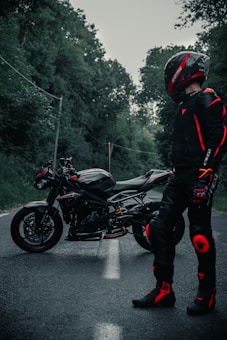 A motorcyclist in full gear, including a helmet and protective suit with red accents, stands near a sleek black sports motorcycle. The scene is set on a quiet, tree-lined road, conveying a sense of adventure and solitude.