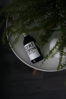 A bottle labeled 'cold brew' is placed on a white circular surface with green leaves draping over it. The setting appears to be minimalist and natural, giving a relaxed vibe.
