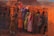 A group of women are engaged in manual labor, carrying and stacking bricks at a brick kiln. They are dressed in colorful traditional clothing, with some wearing headscarves. The surrounding environment is filled with stacks of red bricks, and there is a dusty atmosphere. The warm tones of the setting sun cast an orange hue across the scene.