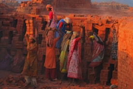 A group of women are engaged in manual labor, carrying and stacking bricks at a brick kiln. They are dressed in colorful traditional clothing, with some wearing headscarves. The surrounding environment is filled with stacks of red bricks, and there is a dusty atmosphere. The warm tones of the setting sun cast an orange hue across the scene.