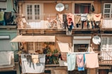 Multiple apartments with balconies display an array of colorful clothes hanging to dry. Two satellite dishes are attached to the building, and there are potted plants on the balconies, creating a rustic, lived-in atmosphere.