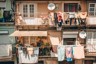 Multiple apartments with balconies display an array of colorful clothes hanging to dry. Two satellite dishes are attached to the building, and there are potted plants on the balconies, creating a rustic, lived-in atmosphere.