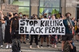 A group of people, predominantly wearing face masks, are gathered in a public space holding signs advocating against racism. The central focus is a large banner with the message 'Stop Killing Black People'. Another sign reads 'Sta op tegen racisme wereldwijd', which translates to 'Stand up against racism worldwide'. The individuals display diverse styles of clothing and appear unified in purpose.