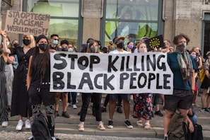 A group of people, predominantly wearing face masks, are gathered in a public space holding signs advocating against racism. The central focus is a large banner with the message 'Stop Killing Black People'. Another sign reads 'Sta op tegen racisme wereldwijd', which translates to 'Stand up against racism worldwide'. The individuals display diverse styles of clothing and appear unified in purpose.