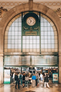An ornate clock with Roman numerals is centered above an arched entrance in a bustling train station. Below the clock, there are digital timetables displaying arrivals and departures. A group of people stand in the foreground, some looking up at the information board.