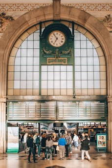 An ornate clock with Roman numerals is centered above an arched entrance in a bustling train station. Below the clock, there are digital timetables displaying arrivals and departures. A group of people stand in the foreground, some looking up at the information board.