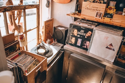 A cozy record store corner featuring a collection of vinyl records and a record player. Sunlight streams through a rustic window illuminating various items including albums with recognizable covers, a small speaker, and a cocktails menu. Shelves and bins are filled with records, creating a nostalgic atmosphere.