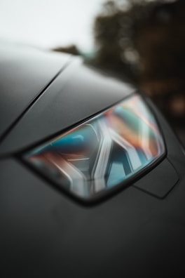 A close-up of a car headlight featuring a sleek design with geometric patterns. The background is blurred, emphasizing the modern and stylish appearance of the car.