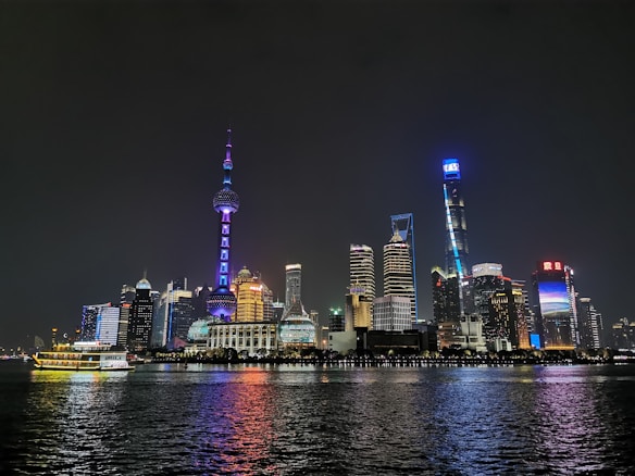 A nighttime cityscape featuring a skyline of illuminated skyscrapers reflecting off a calm body of water. The architecture displays a mix of modern high-rise buildings with vibrant lighting, including a distinctive tower with a spherical design. The scene captures the urban ambiance against a dark sky.