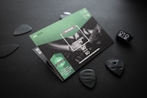 A set of guitar picks and a promotional flyer arranged on a dark surface. The flyer features a partial image of a person playing a guitar. The text 'ECO-BLACK' and 'The new generation of guitar picks' is visible, emphasizing sustainability. Several black guitar picks are scattered around the flyer.