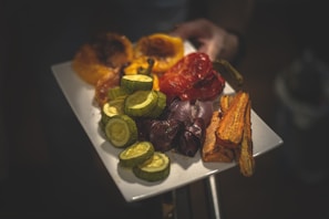 A white rectangular plate holds a variety of roasted vegetables including yellow bell peppers, red bell peppers, sliced zucchini, purple onions, whole mushrooms, and sliced carrots. The vegetables appear to be well cooked and arranged neatly on the plate. A hand is holding the plate.