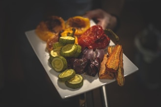 A white rectangular plate holds a variety of roasted vegetables including yellow bell peppers, red bell peppers, sliced zucchini, purple onions, whole mushrooms, and sliced carrots. The vegetables appear to be well cooked and arranged neatly on the plate. A hand is holding the plate.