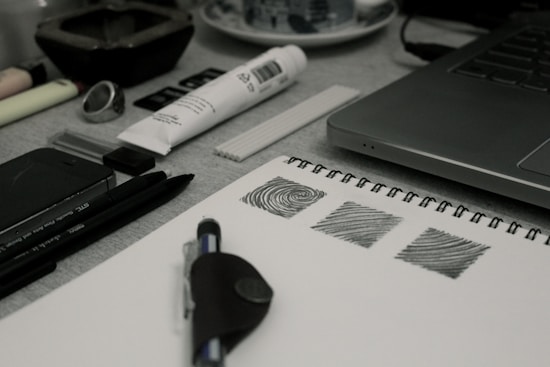 A creative workspace featuring a laptop, a sketchbook with drawn patterns, pens, a marker, a tube of paint, and various art supplies. The items are arranged neatly on a desk, suggesting an environment conducive to art and design.