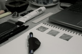 A creative workspace featuring a laptop, a sketchbook with drawn patterns, pens, a marker, a tube of paint, and various art supplies. The items are arranged neatly on a desk, suggesting an environment conducive to art and design.