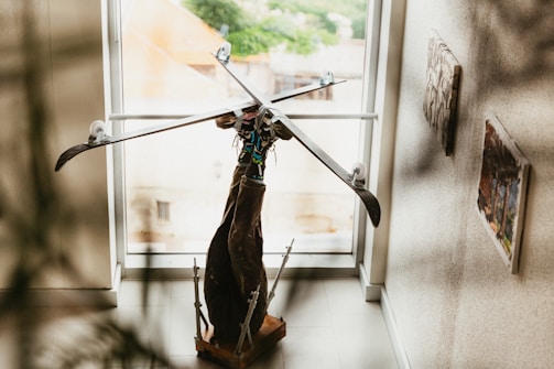 A creative sculpture made from various materials is placed near a large window. The sculpture is constructed with ski poles forming a star-like shape at the top and appears to be supported by wooden elements. Artistic paintings are hung on the walls surrounding the sculpture, and natural light filters through the window, casting shadows across the floor.