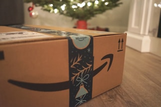 A cardboard box with Amazon branding sits on a wooden floor, with festive blue decorative tape featuring holiday motifs. In the background, a blurred image of a Christmas tree illuminated with lights can be seen, along with a few red and silver ornaments.