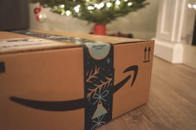A cardboard box with Amazon branding sits on a wooden floor, with festive blue decorative tape featuring holiday motifs. In the background, a blurred image of a Christmas tree illuminated with lights can be seen, along with a few red and silver ornaments.